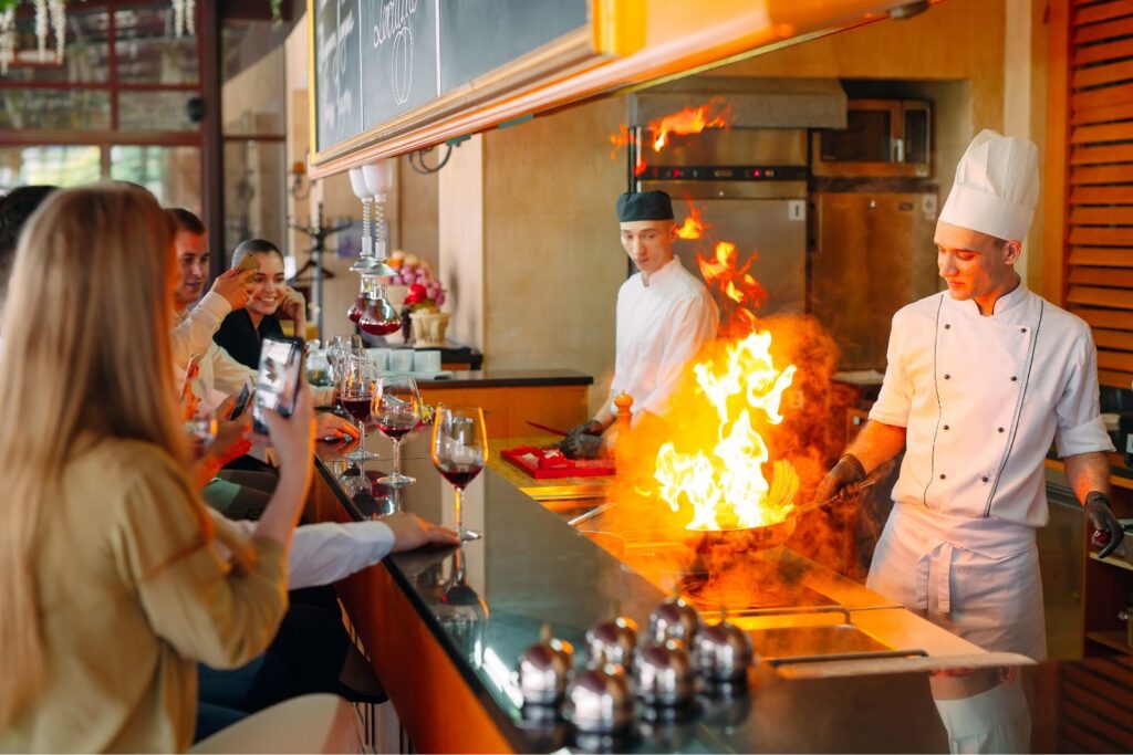Ein Koch flambiert ein Gericht in einer offenen Küche mit großer Flamme, während mehrere Gäste an der Theke sitzen, Wein trinken und mit ihren Smartphones filmen.