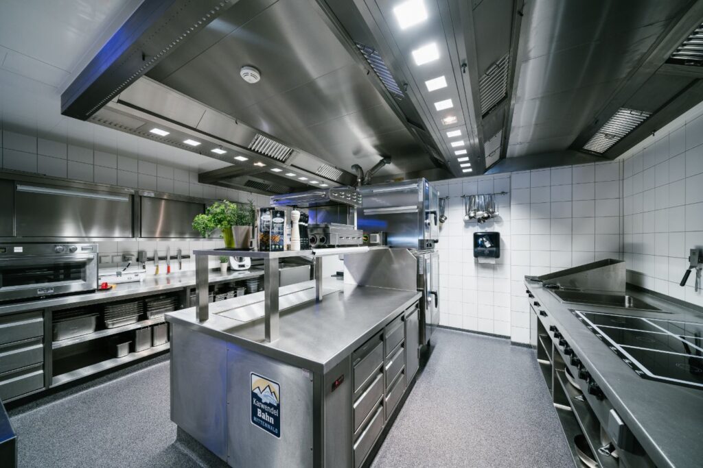 Weitwinkelaufnahme einer Gastroküche mit 2 Theken links und rechts sowie in der Mitte. Oben mittig sieht man die Beleuchtung.